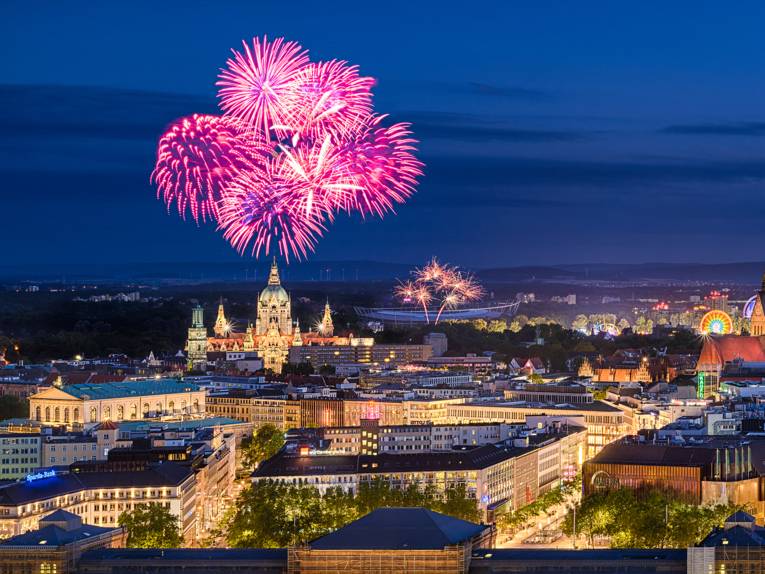 Beleuchtete Skyline von Hannover bei Nacht mit Feuerwerk über dem Neuen Rathaus und bunten Lichtern eines Volksfests.